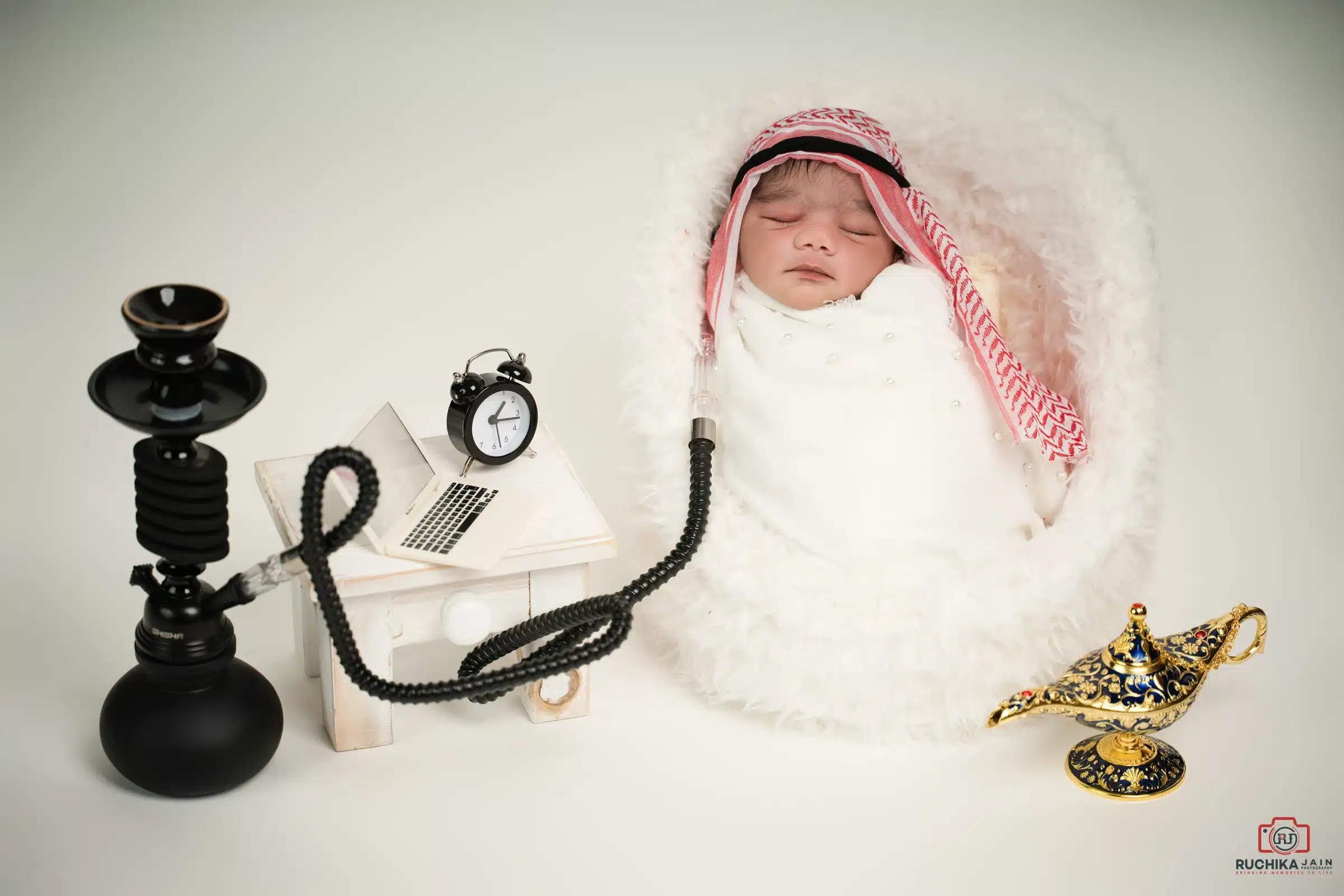 Sleeping newborn wrapped in white fabric, styled with a traditional headscarf and surrounded by playful themed props.